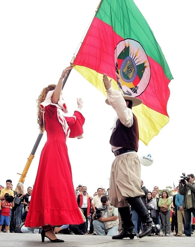 O Peão e a Prenda dançando com a bandeira do Estado do Rio Grande do Sul ao fundo.jpg